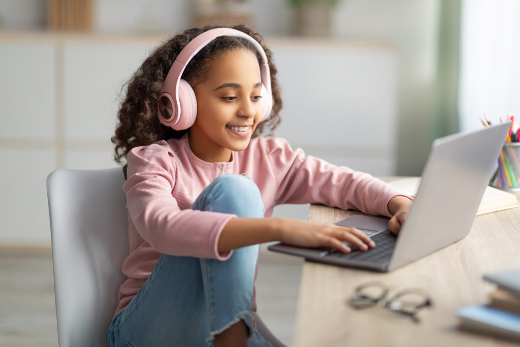Online chat. Happy teenage girl using laptop, wearing wireless headphones, relaxing sitting at desk at home. Young user browsing internet or listening course, typing on keyboard