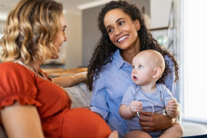 Happy friends with child relaxing on couch at home. Mixed race woman with expecting mother talking and playing with baby. Pregnant lesbian mothers sitting on sofa and smiling with infant