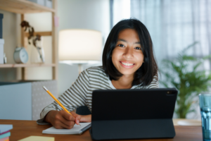 Young girl looking forward and smiling. She is on a tablet and writing in a notebook with a pencil 