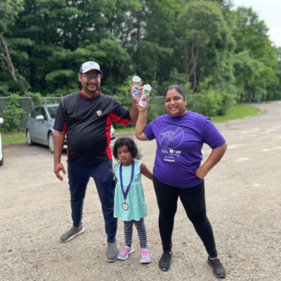 2 smiling parents holding All 4 Her 5k water bottles and a child wearing an All 4 Her 5k medal