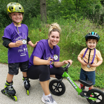 smiling mom and 2 kids wearing All 4 Her 5k medals. Both children are wearing helmets, one is wearing rollerblades and one has a bike