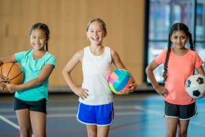 Three girls standing in a gym holding a basketball, a volleyball and a soccer ball.