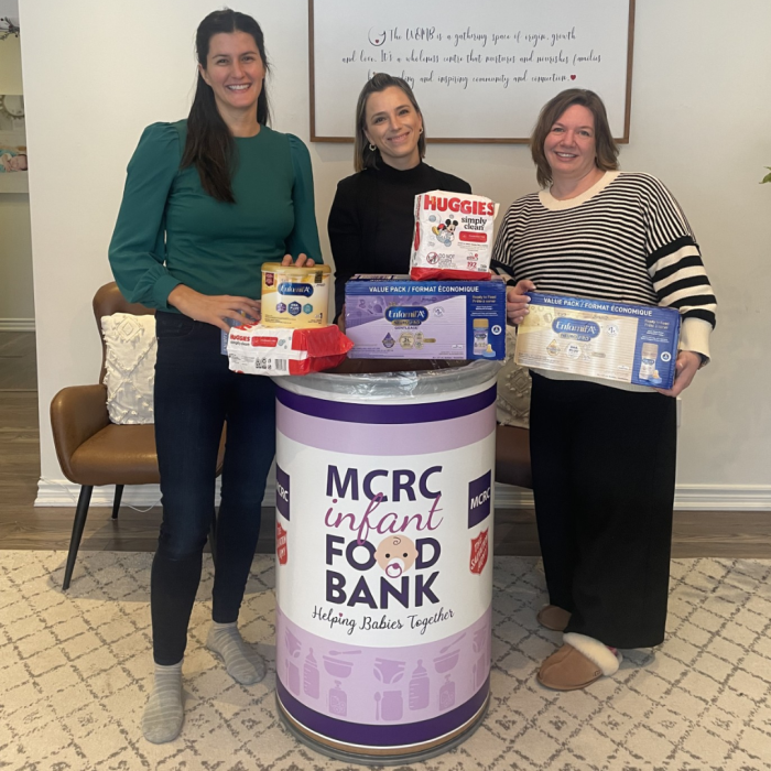 Thursday Jan 2nd (2) 3 women smiling and holding donations of baby items near a donation bin that says MCRC Infant Food Bank- Helping Babies Together