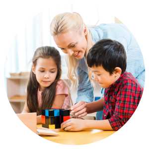 a woman leaning over a table that 2 children are sitting at, guiding them with an activity