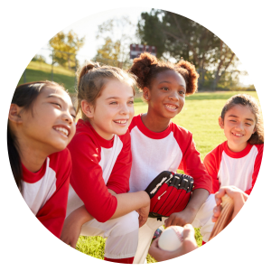 four young girls in baseball shirts and gloves kneeling on the grass smiling