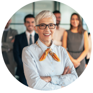 A woman with short gray hair and glasses smiles at the camera, arms crossed, with a diverse group of colleagues blurred in the background.